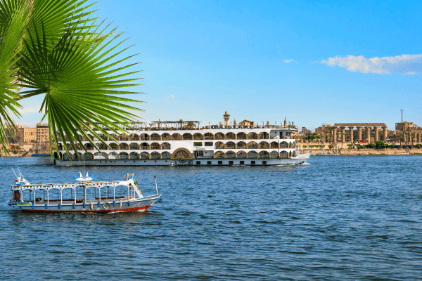 View of a traditional boat and a large cruise ship on the Nile River in Egypt, framed by palm leaves.