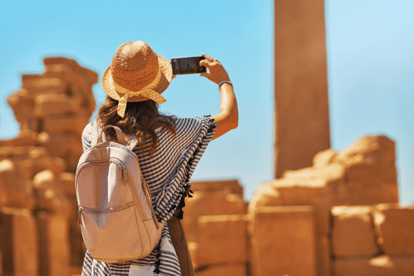 Traveler, traveling with Discovery Tours Egypt, taking a photo of ancient ruins in Egypt with a smartphone, wearing a straw hat, capturing Egypt’s rich history.