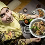 Woman presenting traditional dish, smiling in kitchen.