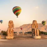 Hot air balloons over ancient Egyptian statues at sunrise.