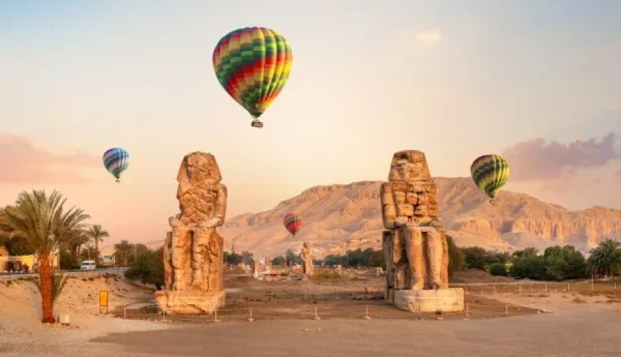 Hot air balloons over ancient Egyptian statues at sunrise.