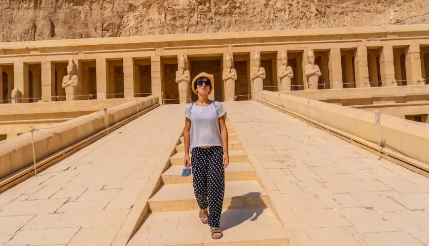 Visitor walking towards the Hatshepsut Temple in Egypt with ancient statues in the background.