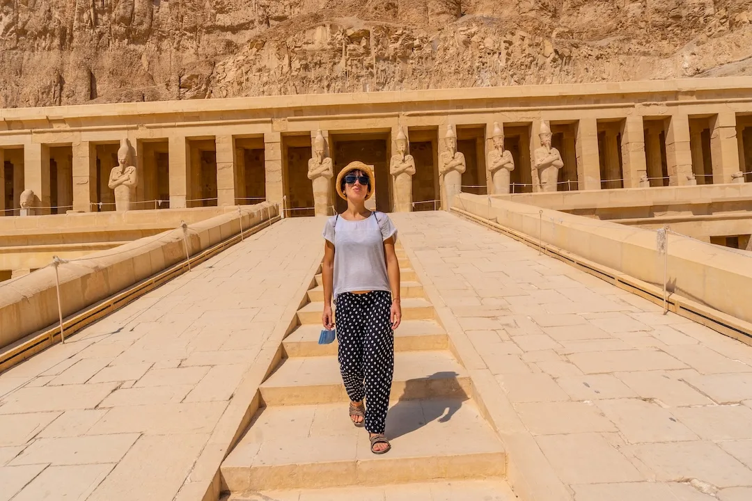 Visitor walking towards the Hatshepsut Temple in Egypt with ancient statues in the background.