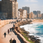 Seaside walkway with people and dilapidated buildings.