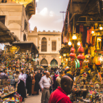 Bustling market street at dusk, colourful stalls and shoppers.